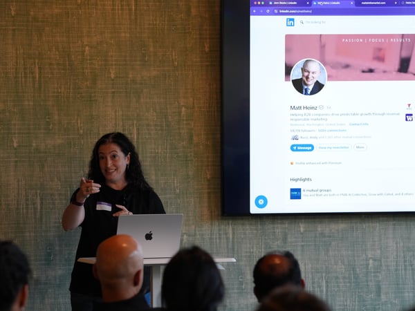 A woman stands at a podium with a laptop, presenting to an audience while a large screen displays a LinkedIn profile behind her.