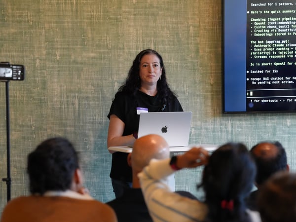 A woman stands at a podium presenting to an audience with a laptop and a large screen displaying text behind her.