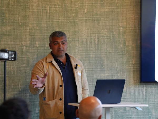 A man in a tan jacket gestures while giving a presentation next to a laptop.