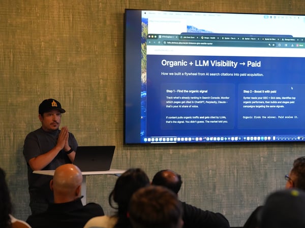 A speaker stands beside a laptop, presenting to an audience in front of a large screen displaying slides about digital marketing and LLM visibility.