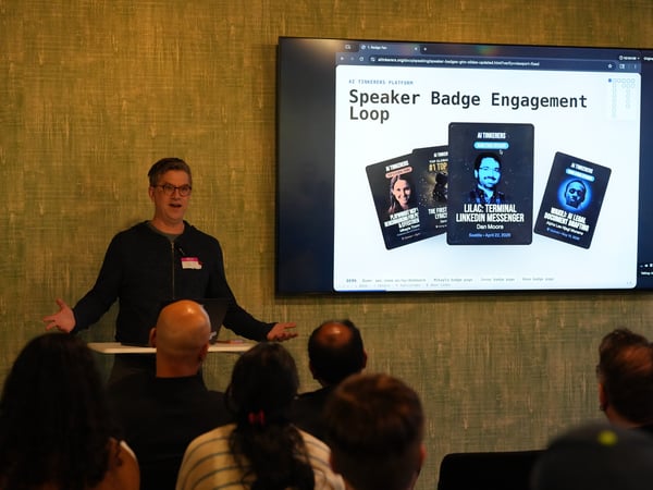 A man stands at a podium presenting to an audience. Behind him, a large screen displays a slide titled 'Speaker Badge Engagement Loop' with examples of digital badges.