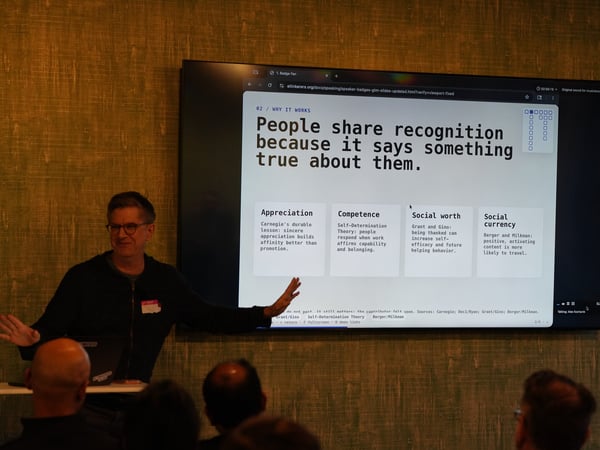 A presenter stands next to a large display screen showing a slide about recognition and social worth to a seated audience.