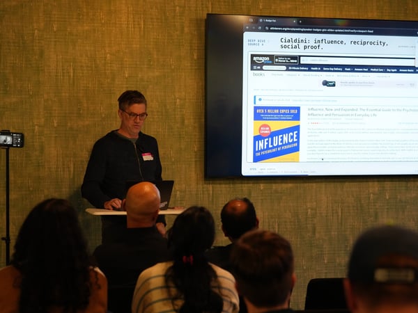 A presenter stands at a small podium next to a large screen displaying a webpage about the book 'Influence' by Robert Cialdini, while an audience watches from the foreground.