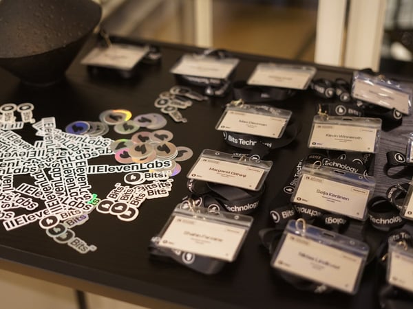 A close-up photograph displays various event materials laid out on a dark table, including numerous name badges attached to lanyards featuring the 'Bits Technology' logo and piles of 'ElevenLabs' and 'Bits' stickers.