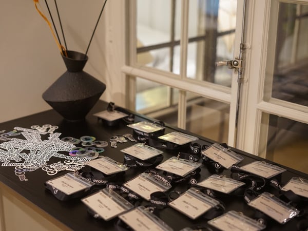 A close-up photograph of a registration table at an event, displaying numerous name badges attached to black lanyards, a pile of 'ElevenLabs' and 'Bits' stickers, and a black decorative vase, set against a large window.