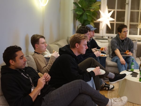 A candid indoor photograph shows five young men sitting closely together on a light-colored sofa, eating pizza and looking toward an unseen screen or focal point.