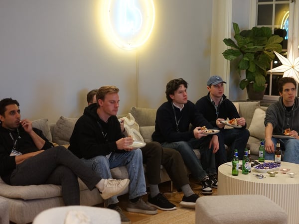 A candid photograph captures a group of six young men sitting closely together on a large gray sofa in a modern indoor lounge, eating snacks and looking intently toward an unseen focal point off-camera. The room is decorated with a distinctive neon light fixture and a large potted plant.