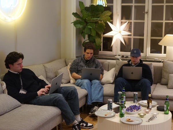Three young men are sitting on a light-colored sofa, intently working on their laptops in a dimly lit, cozy indoor setting. A round coffee table in the foreground holds empty plates and several bottles and cans of beer, suggesting a late-night work session.