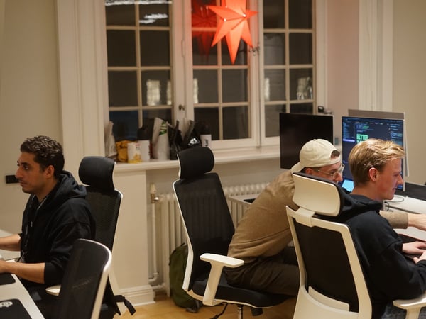 Three young men are working at computer desks in a dimly lit office setting, with two of them leaning in to collaborate on a laptop screen.