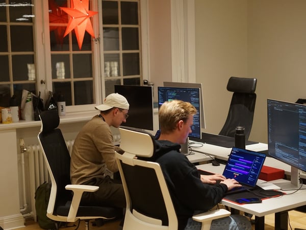 Two young men are sitting at modern office desks, deeply focused on working on their laptops and external computer monitors in a dimly lit office space.