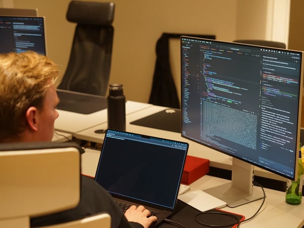 A candid photograph shows a young person with blond hair working intensely on a computer setup featuring a laptop and a large external monitor displaying lines of code in a dark theme.