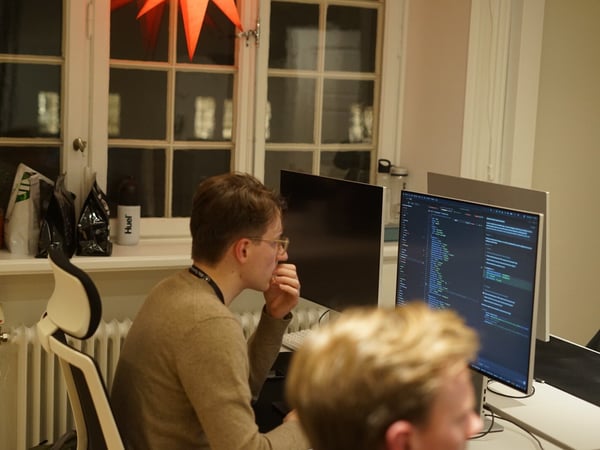 A low-light candid photograph showing two people working in an office setting, with the main subject viewed in profile intently looking at dual computer monitors displaying code.