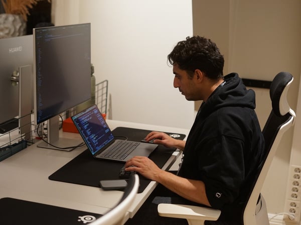 A man wearing a black hoodie sits at a white desk, intensely focused on working on a laptop connected to two large external monitors displaying lines of code in a dimly lit indoor setting.