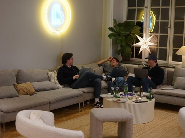 Three young men are casually sitting on a large gray sectional sofa indoors, laughing and engaging with laptops, surrounded by snacks and drinks on a central coffee table, with a neon 'b' sign glowing on the wall.