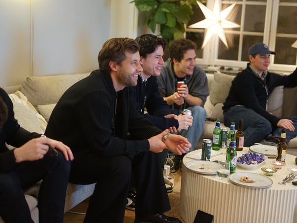 Five young men are gathered on a light-colored couch in a cozy indoor setting, laughing and watching something off-camera while enjoying drinks and snacks placed on a ribbed white coffee table.