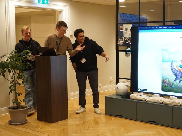 Three young men are gathered around a wooden podium and a large digital display screen in an office setting, appearing to give a presentation or demo of digital content.
