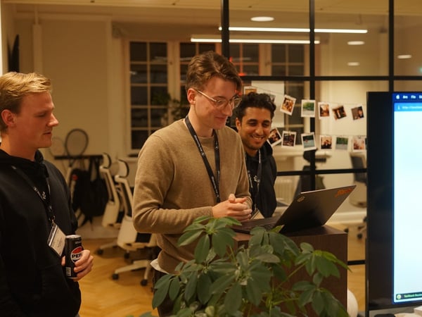 A candid photograph captures three young men gathered around a laptop and a large display screen in a modern office environment, suggesting a casual presentation or technical discussion.