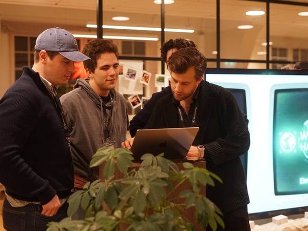 Four men are gathered around a laptop on a small podium, collaborating or reviewing work in an indoor setting, likely during a hackathon event.