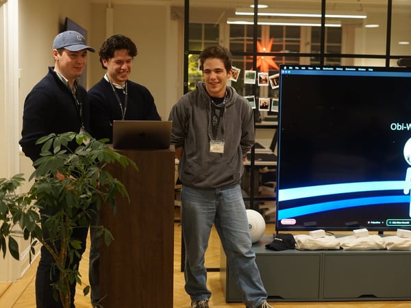 Three young men stand around a wooden podium with a laptop, participating in an indoor event or presentation while a large screen displays a graphic featuring a figure labeled "Obi-Wan Kenobi."