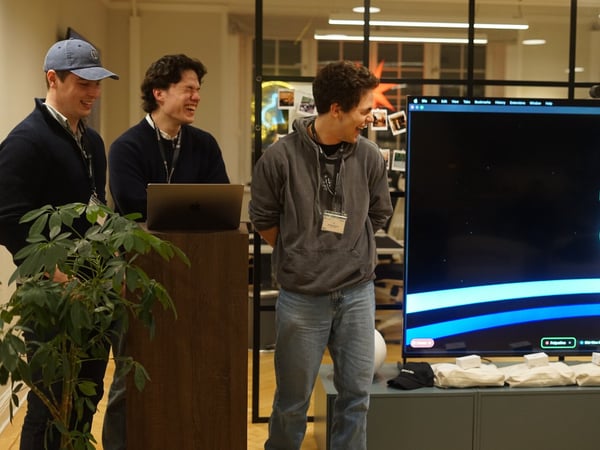 Three young men stand indoors in an office setting, laughing while looking at a large monitor that displays the name "Palpatine" above a simple character graphic, suggesting a presentation or demo is taking place.
