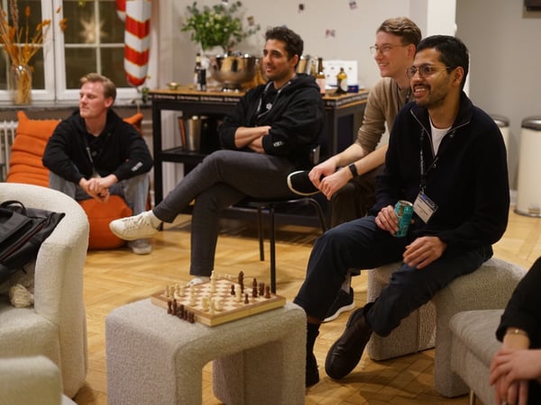 A candid photo capturing a group of men socializing in a relaxed indoor setting, possibly a lounge or office social event, with a small wooden chessboard visible on a low ottoman in the foreground.