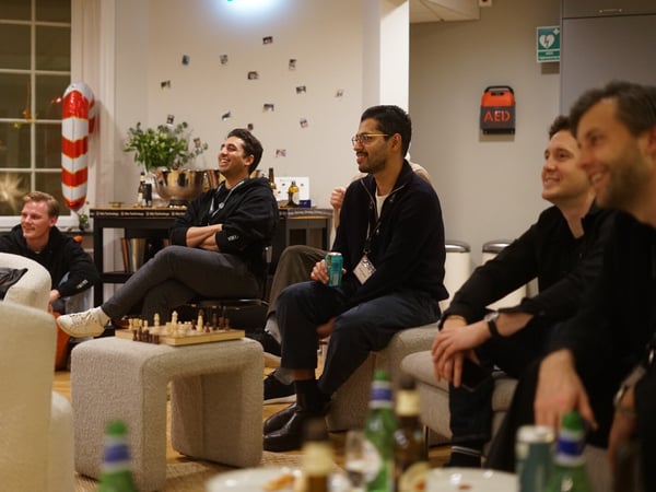 A candid photograph captures a group of men sitting in a relaxed indoor setting, smiling and looking toward an unseen focal point off-camera, possibly during a presentation or viewing party. A small wooden chessboard rests on a fuzzy stool in the foreground.