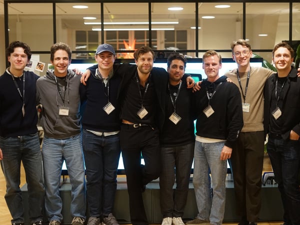 A group portrait of seven young men standing shoulder-to-shoulder indoors, smiling at the camera. They are all wearing casual attire, including jeans and sweaters, and have lanyards with ID badges around their necks, suggesting they are attending a professional event.