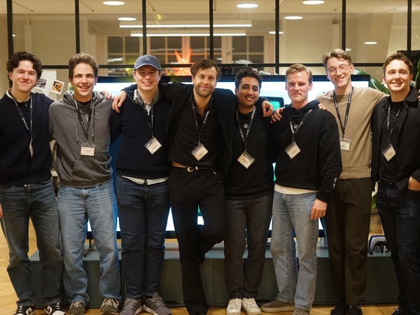 A group portrait of eight young men standing closely together, smiling for the camera in an indoor setting, likely a corporate event or office space. They are all wearing casual clothing and event badges around their necks.