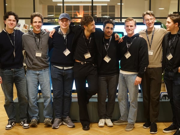 A group of eight young men, dressed casually and wearing lanyards, stand shoulder-to-shoulder posing for a photo indoors, likely in a modern office or event space.