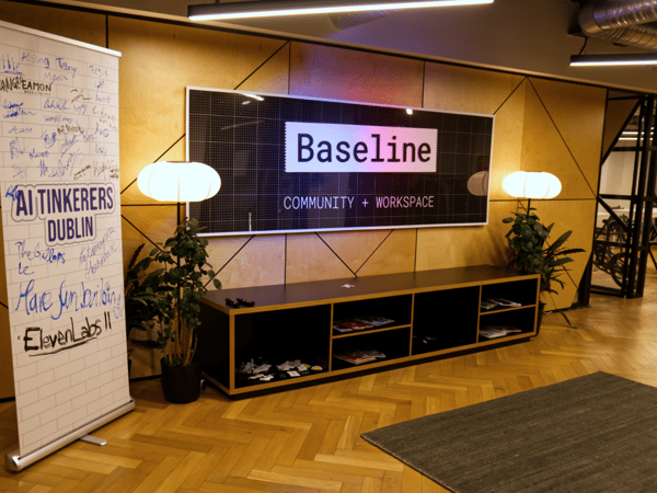 A photograph of a modern workspace interior featuring a wooden wall with geometric paneling, above a low black shelving unit and a parquet floor. A large backlit sign displays the text "Baseline COMMUNITY + WORKSPACE," and a pull-up banner covered in handwritten signatures and messages stands to the left.