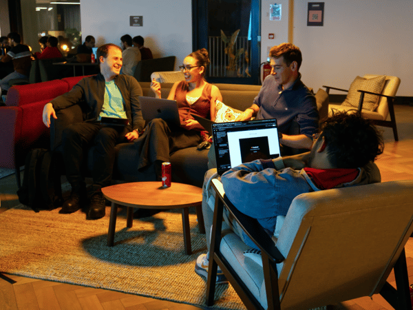 Four individuals are gathered in a modern, dimly lit lounge area, sitting on comfortable furniture and collaborating or working on open laptops.