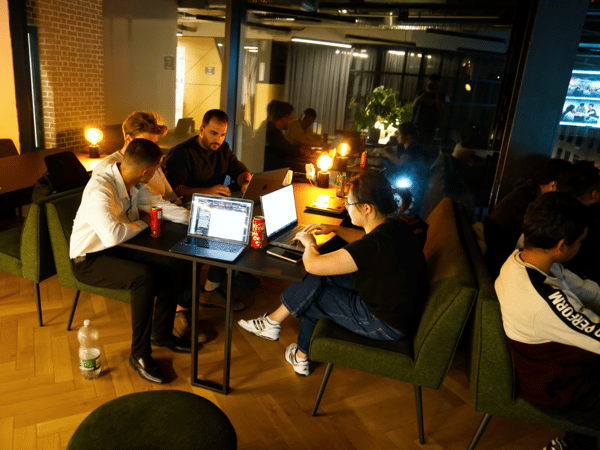 Four people are seated around a small table, intently working on laptops in a dimly lit, modern indoor setting, possibly during a late-night coding session or hackathon.
