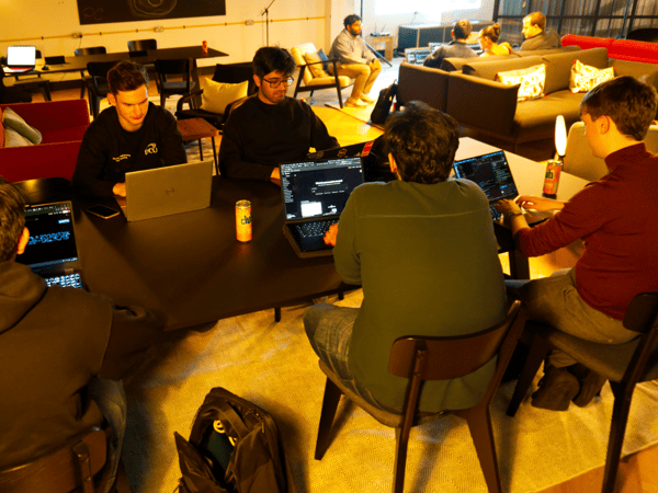 A candid photograph captures a group of young adults intensely working on laptops around a dark table in a dimly lit, casual indoor lounge or co-working space, while other participants relax on couches in the background.