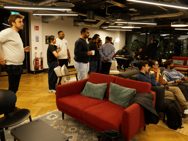 A candid photograph captures a diverse group of people gathered in a modern, stylish office lounge area, with some standing and others seated on couches watching or listening to an informal discussion or presentation.