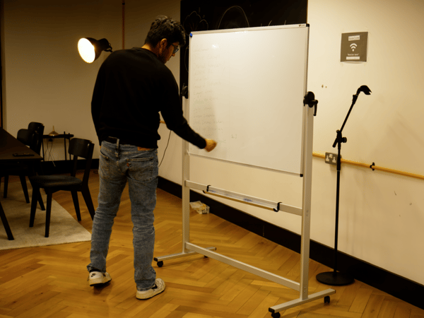 A photograph captures a man, viewed from the back, writing notes on a mobile whiteboard in a dimly lit indoor space with wooden flooring.