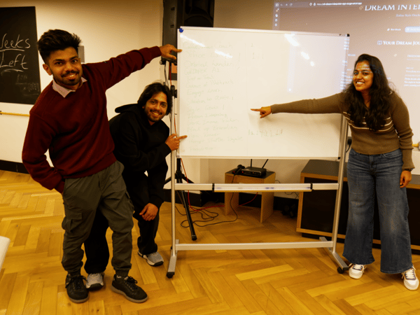 A group of three people (two men and one woman) stand smiling around a whiteboard, pointing at the handwritten list, while a projection screen displaying a website titled "DREAM INTERPRETER" is visible behind them.