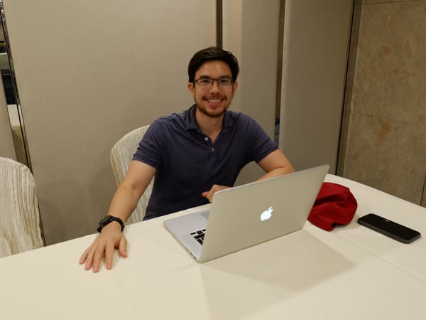 A man sits at a white table with an open laptop, looking towards the camera and smiling.