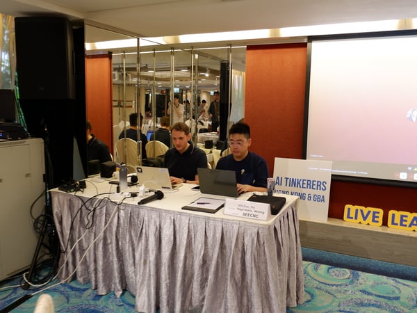 Two men are working on laptops at a table in a conference room, which features a large projection screen and signs for 'AI Tinkerers Hong Kong & GBA'.