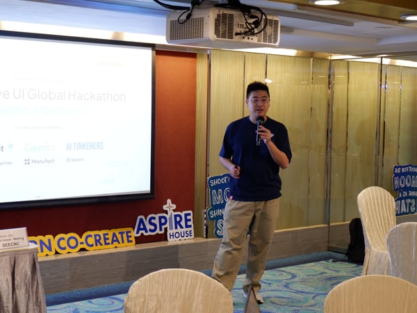 A young man is giving a presentation at the Generative UI Global Hackathon, standing in front of a projector screen with event branding.