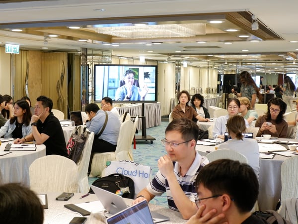 A group of people are seated at round tables in a large conference hall, attending a presentation shown on a large screen.