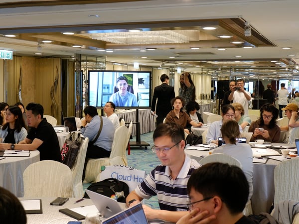 A group of people are gathered in a large indoor hall for a workshop or conference, sitting at round tables with laptops while a speaker is displayed on a large screen.