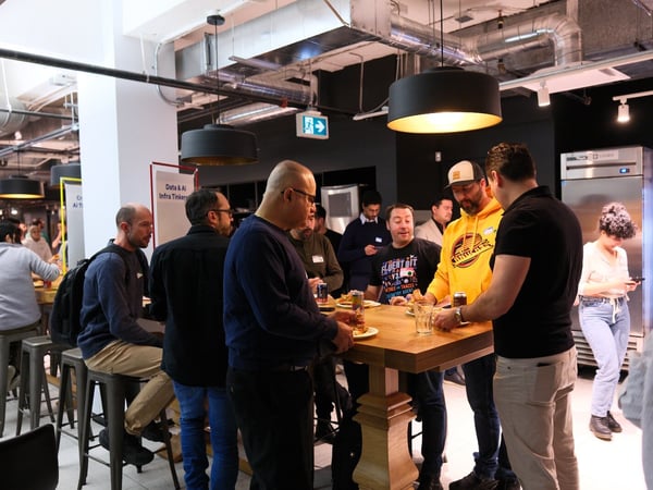 A candid photograph captures a group of people socializing and networking around a high wooden table in a modern indoor venue with industrial architectural elements.