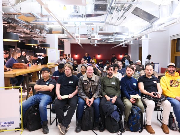 A wide-angle indoor photo shows a large group of attendees gathered for an event, with a front row of nine men seated and facing the camera, while others mill about or sit at communal tables in the background.