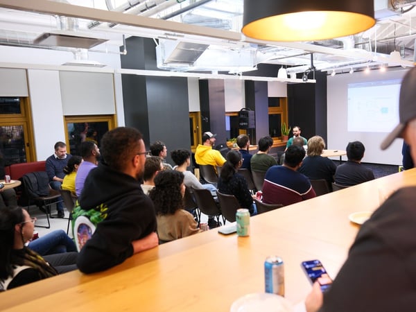 A candid photograph captures a diverse audience seated in a modern indoor venue, watching a presenter stand near a large projection screen displaying a technical diagram or interface.