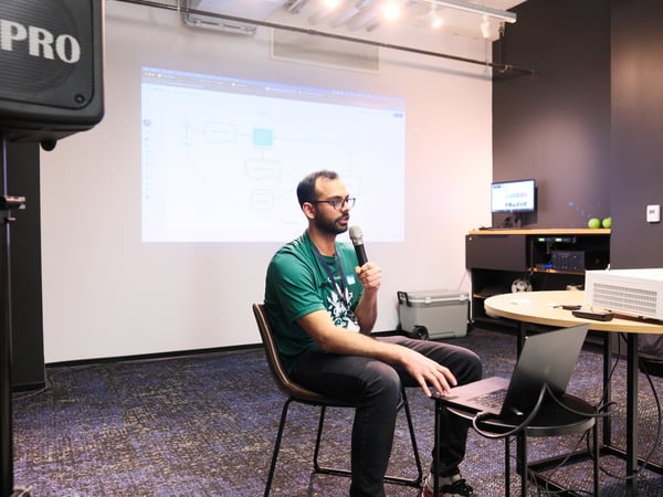 A man wearing glasses and a green shirt sits on a chair, holding a microphone and speaking during a technical presentation in an indoor event space.