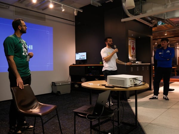 A candid photograph taken indoors showing three men participating in a presentation or meeting, with one man speaking into a microphone near a projector and a large blue slide visible on the wall.