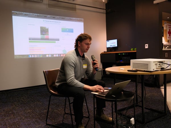 A man wearing a grey sweater sits on a chair, holding a microphone and operating a laptop while presenting a data analysis application projected onto a large screen in a modern, dimly lit indoor space.