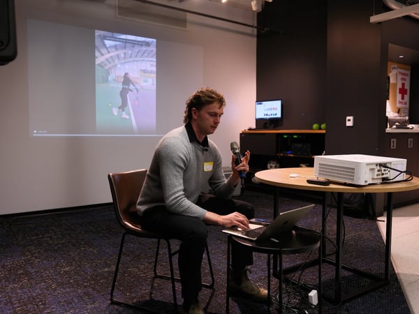A man is seated on a chair, holding a microphone and using a laptop during an indoor presentation or talk, with a video projection of a person playing tennis visible on the wall behind him.