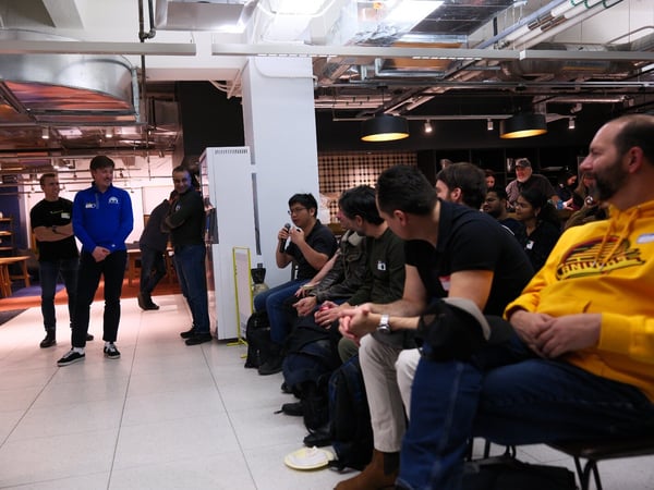 A candid indoor photograph captures a group of attendees seated and listening during an event, while a few organizers stand nearby in a modern industrial-style space. One seated man in the foreground holds a microphone, suggesting a Q&A session or discussion.