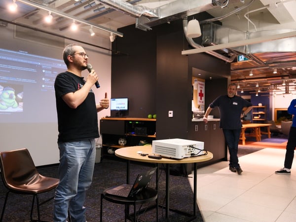 A man wearing glasses and a black shirt is giving an indoor presentation, holding a microphone next to a projection screen displaying text and a cartoon image of two animals.